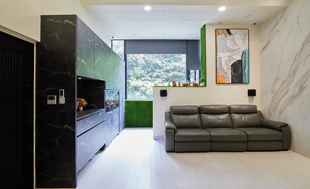 
客厅
Living Room

客厅窗畔预留一处储藏空间，供屋主一家收纳日常用品；沙发旁壁面藉特殊漆仿拟石材肌理，与对侧的玄色石纹柜体相呼应，邻接电视牆则设计为灰色，结合雷射电视的投影幕提升视觉层次感，上方壁面嵌入大片灰玻，不仅能从二楼看顾孩童，也兼顾私领域隐私。

We set up a storage nook that ingeniously incorporates the living room window side, providing homeowners with a convenient space for stashing everyday essentials. The wall adjacent to the sofa features a unique finish replicates the natural stone texture, harmonizing with the opposite dark stone-patterned cabinet. Adjacent to this, the TV wall adopts a subtle gray tone and is perfectly complemented by a projector screen that enhances the visual depth. Above, a large expanse of gray-tinted glass has been integrated into the wall, offering a dual purpose: it allows parents to keep an eye on their children from the second floor while ensuring the privacy of personal spaces.
