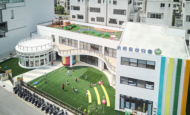 本文由OPEN编辑部撰写

设计概念

整齐林立的宁静住宅区中，一幢多彩明亮的建筑吸引众人目光，斑斓色彩间以圆润曲弧细腻缀点，勾勒柔和舒缓的自在氛围，庭院内，幼童于宽阔无碍的绿地中恣意奔跑、尽情嬉戏，灌注活泼轻快的童真气息，一张张展露灿烂笑颜的脸孔，代表孩童们共度的美好时光，而留存在心中无可取代的快乐回忆，也将化为未来健康成长的养分。

考量学龄前儿童正处好奇心旺盛的阶段，D.H.I.A黄静文室内设计 黄静文 设计总监 以多变的几何、流线造型为主要设计语彙，不仅和周遭方正建物形成强烈对比，也成就层次感丰富的趣味空间。园区内立面、天壁粉饰上鲜明的绿、黄、蓝、红，酝酿俏皮生动的童趣感，同时呼应「处处皆可学」的精神，鼓励幼儿认识、探寻未知颜色，增进想像力及创造力。

设计手法

多元区域 陪伴成长

本案将建物设于基地后侧，前方规划铺设人工草坪的宽敞广场，与配有跑道、小山丘、跷跷板和凉亭的顶楼空间，供孩子们进行各式户外运动，结合位在三楼室内的益智游戏室，既可因应台湾多雨的气候，也予以幼童更多样化的活动选择。

活用天光 自适舒心

全案廊道设有对外落地门窗，创造敞亮的阳光走廊，而教室壁面的圆弧、几何造型窗及嵌入玻璃的门片，也引入大量户外天光，搭配清新鲜明的色彩，形构明亮舒适的学习场域。

缜密思量 完善机能

黄总监于各地坪交接处规划平缓的斜坡，辅以可容纳多人的电梯和无障碍厕所，给予行动不便者友善的环境，另将办公室和厨房规划于一楼教学区外的圆形空间，提供教职员宁静的工作场所，比邻处则设置保健室，除了方便师长就近照看孩童，独立的区域也能确保休息品质，避免与他人接触传染，维护学童们的健康安全。
