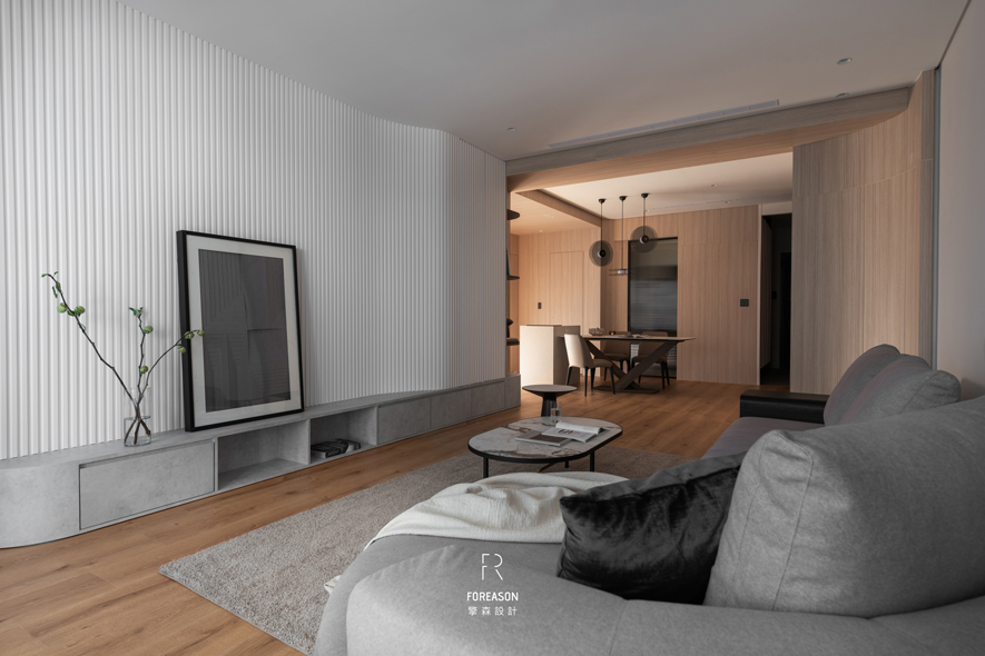 
客厅——留白中的温柔对话

电视牆以细緻的直条纹肌理为底，简约的灰白色调让视觉得以舒缓。灰色L型沙发如云朵般柔软地坐落于木地板上，搭配深浅不一的抱枕与毛毯，邀请家人随时陷入舒适的拥抱。弧形落地灯优雅探身而来，在阅读时光裡映照恰到好处的柔光；两张圆形茶几错致摆放，承载随手翻阅的书籍、温热的茶杯，和午后慵懒的阳光。没有多馀的装饰，这些不经意的细节让空间在留白中自成风景，于从容中展现真实的生活质感。
