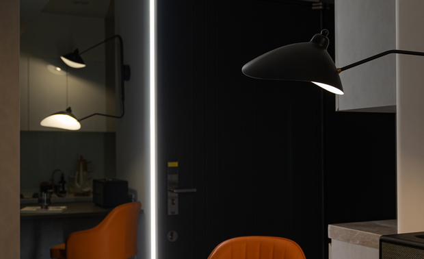 
餐厨区——开放格局 凝聚情感的轻食天地

步入室内，中岛吧檯作为空间的核心映入眼帘，串联厨房与客厅的日常动线，成为整个空间交织的枢纽。灰褐色檯面沉稳内敛，与前方白色橱柜的明淨俐落相互呼应，共同勾勒出简约的料理场域；旁侧，一张橘色高脚椅为这片宁静基调注入活泼生命力，而在黑色造型吊灯下，復古音响等摆件点缀其间，每一处细节都为生活留下表达的馀地。入夜后落座单椅，后方镜面映照着光影流转，整个角落彷彿与世隔绝，成就独属自己的静谧时刻。
