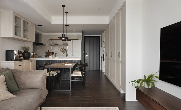 
玄关、餐厨区──沉敛迎序 静好入室

推门而入，黑色直条的玄关门扉率先定下静谧的序曲，两侧浅木柜体整齐归置零散物件，一深一浅相映，不疾不徐地勾勒出归家的第一道仪式感。视线顺着廊道延伸，餐厨区随即以开放之姿迎面展开——餐桌檯面藉浓郁木调安稳定锚，一旁石纹中岛白灰檯面则轻柔呼应，搭配黑色铁件餐椅及头顶两盏俐落的圆盘吊灯，于暖冷调的交会处拿捏出恰好的生活温度。

中岛后方厨房壁面覆上浅灰涂料铺叙质朴肌理，开放层架上随兴摆放的器皿、咖啡机，不刻意却成一幅有烟火气的静物小景，悄悄透露由此烹茶备餐、对坐閒谈的寻常好时光。
