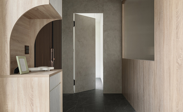 
玄关——弧光引渡 归心之境

踏入门扉，曲面木作隔屏如月牙般优雅延展，巧妙勾勒出内外之界。窗櫺引入的日光洒落墨玉般的石纹地坪上，形成光律交织的迎宾序曲。弧形木作包樑与玄关柜採用相同浅木质调，从立面延伸至顶塑造连续包覆感，消解原有建筑结构的刚硬转角，犹如安稳的臂弯，静候归人卸下风尘；在这初得安稳的怀抱中，旁侧隔间牆转角的弧形导引柔化空间的转折，更进一步引领从外界喧嚣至完全沉淀内心宁静的步履，在这光线递变的过渡之境，心绪随着曲线的韵律，缓缓沉淀，回归家的怀抱。
