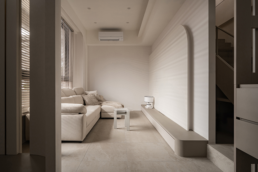 
客厅——温润静谧 推门的第一声轻语

推开门扉，整面暖灰柜体藉由俐落线条安静伫立，转身步入客厅，轻浅色调的白随即舒展开来，牆面循着一道柔缓曲线化解直角生硬，一旁浅色格栅隔屏轻盈地界定廊道进退，在连贯中悄悄梳理动线鬆紧。靠窗一隅，奶油色沙发被窗外引入的日照轻拥，透明茶几映着窗外树影，前方电视柜沿弧牆水平铺展，机能退居幕后，留出大片空白任光影自在描画，拼写安静却有温度的每一天。
