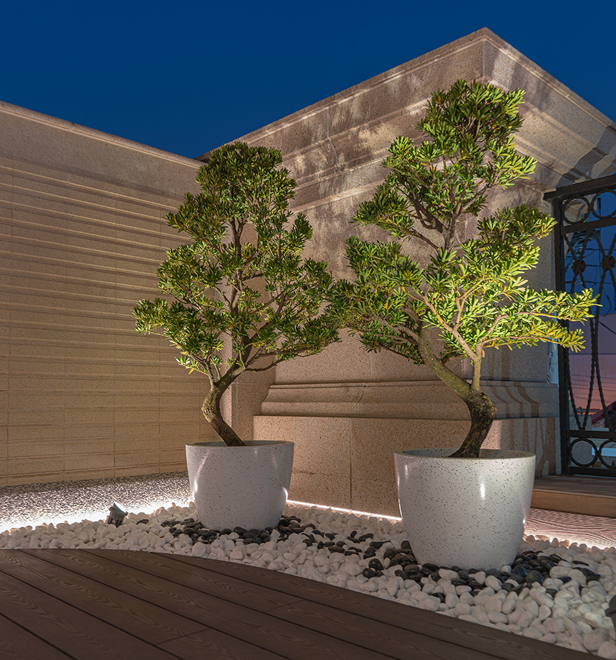 
花圃露台——亲手营造的私密绿洲 灯光植栽刻画夜色风景

夜幕低垂，二楼露台在深蓝夜空的映衬下，展现另一番禅意景致，设计团队与屋主携手，以木质地板铺陈和煦基底，黑白双色卵石交错铺设，配合周围嵌入的间接照明，其光晕温柔勾勒石材、木纹的细腻质地。卵石之上，两颗绿植挺立于洁白陶盆，苍劲枝干与翠绿针叶在夜色中依然盎然生机，透过精心配置的地灯投射，将植栽的优雅身姿映射于牆面，构成一幅流动的光影画作，在有限中创造无限的閒适气氛，成为一家人在忙碌中得以放鬆身心、亲近自然的秘密花园。
