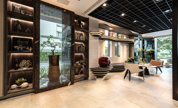 
一楼入口接待、办公区——如何让户外景观和室内设计相互呼应？

在设有大面玻璃窗的结构下，户外与室内元素是否有关联性，会对画面有无一体感产生很大的影响。团队故于此案入门处设置120公分宽的大门，为屋中迎入开阔视野，结合一道融有长虹玻璃和灰玻、向内转折15度的半透明屏风，打造适度遮挡视线的曲弧界面，延续户外水景的流动意象，并兼顾引导动线及引入天然光线、景观的视觉效果。

一旁的办公区进一步将弧型语彙揉入工作机能，依照设计师绘图、办公、收纳等各式需求，彷拟建物外的水池涟漪量身订製大型弧形办公桌，延展桌面尺度也柔化办公家具的量体感，并于四周选置深色柜体搭配俐落灯光，调和出凝鍊柔润流动、沉稳专业的精緻美感。
