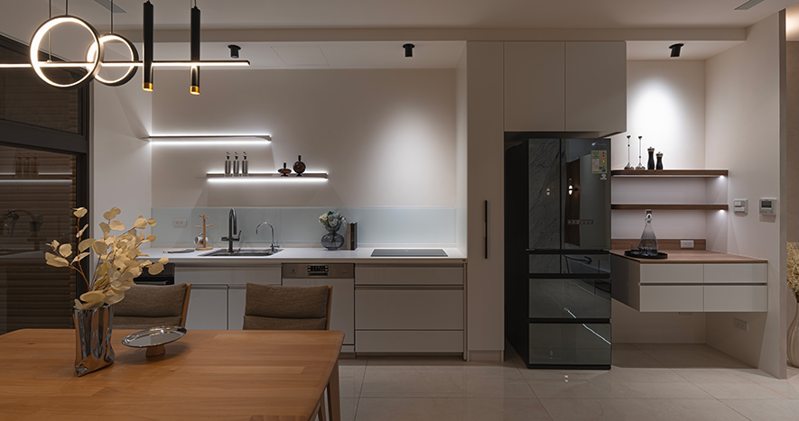 
餐厨区——简淨线条串连生活日常 开放场域凝聚暖意食光

洗练白色系统柜沿牆面一字排开，之中黑色镜面冰箱错落其间，实现恰到好处的色彩对比；牆面上长条灯带为层板投射出柔和光晕，让餐具器皿也成为日常风景。中央实木餐桌以温润质感注入自然气息，围塑一家四口共享美好的核心时光；上方悬挂的环形吊灯，以流畅线条划破空间留白，黑色框架与金色点缀的层次，更为用餐时刻增添一份仪式感。清透白色系与温和中性原木色，在餐厨区中交织清爽明淨的空间表情，而开放格局让烹饪不再是孤单作业，更是串连起家人情感交流的暖心起点。
