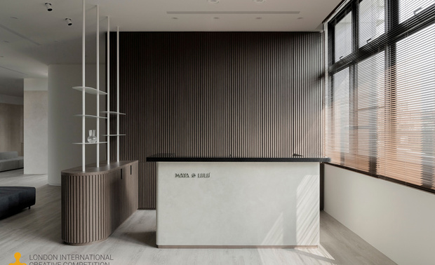 ▲入口柜体以大地色系特殊涂料铺叙沉稳底蕴，迎宾区域廊道则选用灰白色调，营造宁静舒心的过渡场域。接待柜台右侧大面窗引入自然光，与背后格栅式的实木条映照出阴影变化，衬托出自然温润质感。
