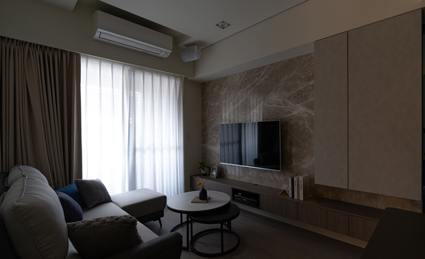 
客厅——光影律动 营造温馨聚会核心

客厅是时光停驻的所在之处，L型沙发与双层圆形茶几围塑出温暖的谈话场域，承载着家人閒聊或独自静思的片刻悠閒。大面积落地窗引入充沛日光，白色纱帘为光线披上轻柔滤镜，随着时序流转，光影在空间中缓缓移动，并搭配灰色布帘的双层调节，使居者能在明暗之间找到最舒适的生活节奏，让日常在细腻的光影变化中流淌；而当夜幕低垂，牆面那盏金色壁灯便如暖阳般为灰白基调增添温柔的注脚。在这份柔和氛围中，电视牆以石材提供一处沉静的视觉锚点，一旁悬浮柜体轻盈俐落，将收纳机能隐于无形，让视线得以纯粹延伸，成就一处卸下疲惫的归心之所。
