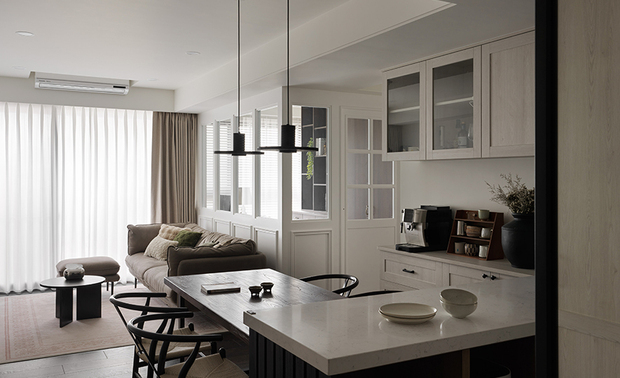 
客厅──轻纱映暖 安然栖心之所

望向客厅，大面落地纱廉筛入柔和天光，为场域披上一层澹澹的晕染。深棕色布沙发低敛而鬆软地倚窗而坐，一旁的圆形茶几同沙发色的圆润矮凳以沉静姿态相伴，悄然守住属于放鬆的那寸馀地。电视牆一侧，悬浮式木质矮柜轻巧贴壁，一盆翠绿蕨叶随意点缀其上，替素淨的浅色基底捎来一丝自然生气，同对向白色壁板、玻璃格扇门的通透光韵形成一场无声的对话——不张扬，却正好将此收拢成一处让人甘愿放下手机、窝进沙发裡好好发呆的柔软角落。
