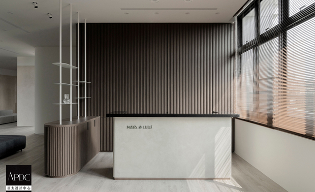 ▲入口柜体以大地色系特殊涂料铺叙沉稳底蕴，迎宾区域廊道则选用灰白色调，营造宁静舒心的过渡场域。接待柜台右侧大面窗引入自然光，与背后格栅式的实木条映照出阴影变化，衬托出自然温润质感。
