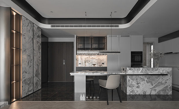 
玄关&餐厨区——石材纹理铺叙入门气度 光影层次勾勒生活质感

设计团队以石材的磅礴纹理为引，于入门处便揭开这方居所的精緻序幕；玄关牆面选用大理石铺陈，其中蕴含的深浅交错肌理，搭配深色柜体的沉穆内敛，奠定静谧的基调。左侧展示柜以金属线条分割层架，内嵌式灯带自上而下透出柔润光晕，让归者甫踏入门扉，便能在光线交织中感受家的温度，营造一处静心的缓冲地带。

步入室内，开阔餐厨区映入眼帘，中岛吧檯以同款大理石延续玄关的设计语彙，厚实的檯面纹理流转有致，既保有场域的独立机能，又形成视觉上的流畅连贯、维繫空间整体的和谐韵律。中岛上方悬挂的水晶灯饰如冰凌般参差错落，晶透晖影洒落于檯面，为料理时光增添几分灵动姿态。一旁，浅灰色调的柜体与天花维持着整体色系的一致性，简洁俐落的线条勾勒纯粹美学风格，而内嵌于牆面的展示柜与背光设计，使收纳与展示在此自然交融，日常餐食准备也因此成为一场赏心悦目的生活仪式。
