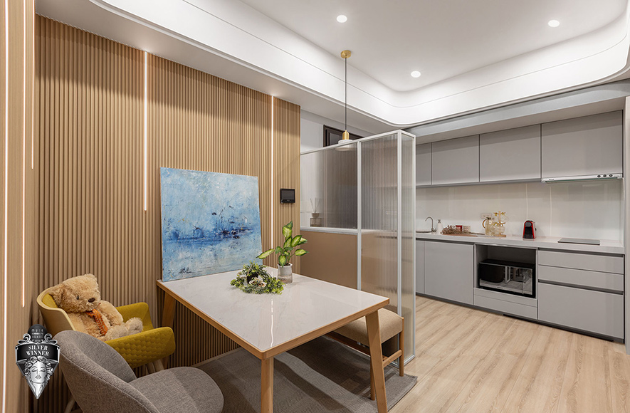 ▲长虹玻璃隔屏以半透光影自然界定玄关领域，保有内外空间的视线缓冲，也为随身物品等预留专属收纳位置。公领域格栅牆面嵌入垂直灯带，光线自细密条纹间溢出，勾勒日系沉静韵律；一桌两椅一凳的精简配置取代传统沙发，使餐区成为可供家人用餐、朋友聚会、工作简报甚至孩童手作的多功能场域。
