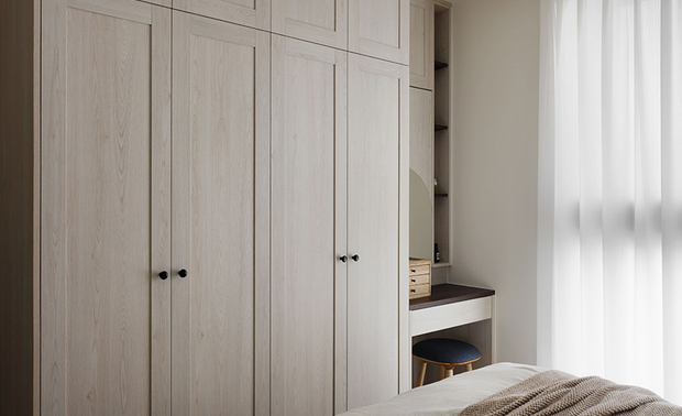 
主卧──暖褐轻覆 归于柔软的日暮时分

一片温润如暮色的灰泥质感壁面，手作般的细腻纹理轻柔包覆整座床头，为卧房铺上一层静谧内敛的底色。床畔两侧，藤编、陶质檯灯各据一方，晕散出如烛火般摇曳的橘黄光晕，同窗边透纱筛入的柔光彼此接应，在白色寝被、驼色针织毯之间晕染出深浅交叠的暖褐色阶。

床尾处，顶天浅木衣柜藉整齐的线脚收齐牆面，靠窗巧妙留出小巧梳妆区，圆弧镜面及木质小抽屉低调素朴地嵌于其间，替日常梳整保留一处幽静而私密的角落。不靠繁复铺陈，只以质地与明暗变幻的层层叠叙，便轻轻接住整天的疲惫。
