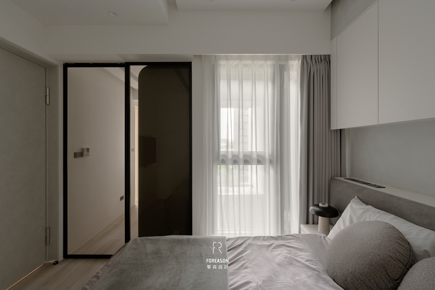 
卧室——灰白之间 沉淀身心疲惫

卧室延续全室的简洁色调，定调浅灰与米白交织出轻柔氛围。床头採用深灰色软包设计，细緻触感托住疲惫身躯，上方的白色吊柜提供充裕收纳，维持立面的纯淨俐落，牆面凹槽设计则保留下方插座使用空间，配合两侧床头柜容纳睡前阅读的书籍、一杯温水，满足居者休憩时的日常所需。窗边双层窗帘展现贴心巧思，白色纱帘过滤晨光成柔和漫射，深灰遮光帘则守护夜晚安眠时光，可随心调节光线明暗及隐私需求，成就让身心沉淀、安然入梦的私密场域。
