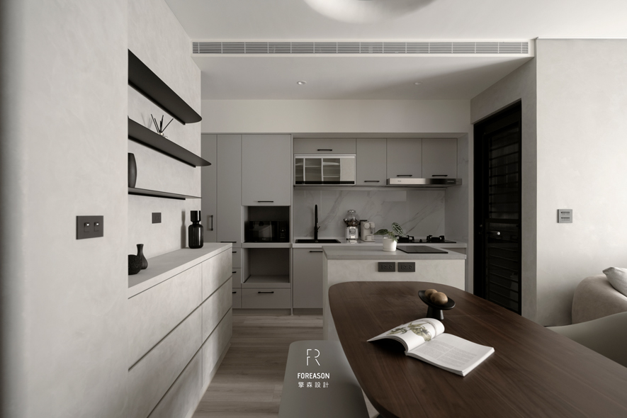 
餐厨区——纯粹立面 定义优雅食光

天花以圆弧内凹搭配隐藏灯带，光线细腻漫溢至整个空间，像是为生活披上一层静谧的滤镜。背牆下方白色柜体採无把手设计，平整立面如同留白画布，上方设置黑色金属层板水平延展，让器物的陈列也成为空间设计的一部分。厨房配置灰色烤漆橱柜和白色檯面，构成简练清爽的视觉效果，中岛深灰石材倒映着光晕，成为料理时的舞台中心及家人围聚閒聊的据点。线条与转折在此重新定义餐厨空间的意义，承载日常裡所有值得被珍视的相聚时光。
