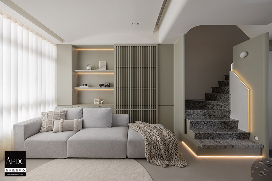 ▲电视牆採用与楼梯踏面相互呼应的纹理铺陈，透过深色调的沉稳质感、如水墨渲染般的自然过渡，平衡黑白两色居多的环境色彩，对向沙发背牆以木作格栅与开放层架自然融合，在线性元素的韵律中创造收纳与展示的双重机能，让生活物件成为空间美学的一部分。
